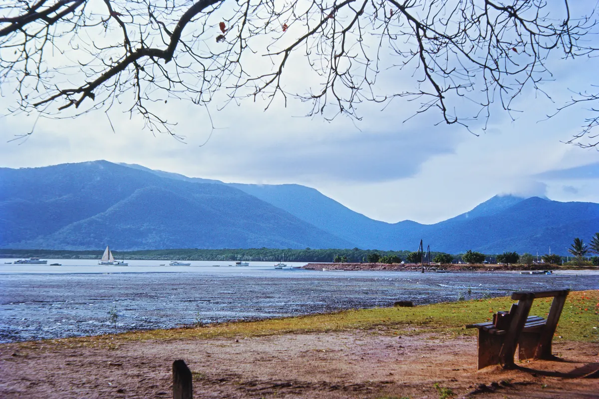 Trinity Inlet's mudflats at low water in front of the hills near Cairns, with a small sailing boat on the far side and a bench in the foreground